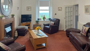 A living room with a fireplace, a television, a leather armchair, a sofa, and a window with a view of the countryside. - Muirston Farmhouse - UK44977 (Cumnock)