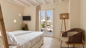 A bedroom with a bed, a television, a wicker chair, and a view of an outdoor pool area. - Villa 'The Blue Diamond' with Sea View, Wi-Fi and Air Conditioning (Mykonos)