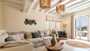 A living room with a sofa, a coffee table, a chair, and a view through a glass door. - Villa 'The Blue Diamond' with Sea View, Wi-Fi and Air Conditioning (Mykonos)