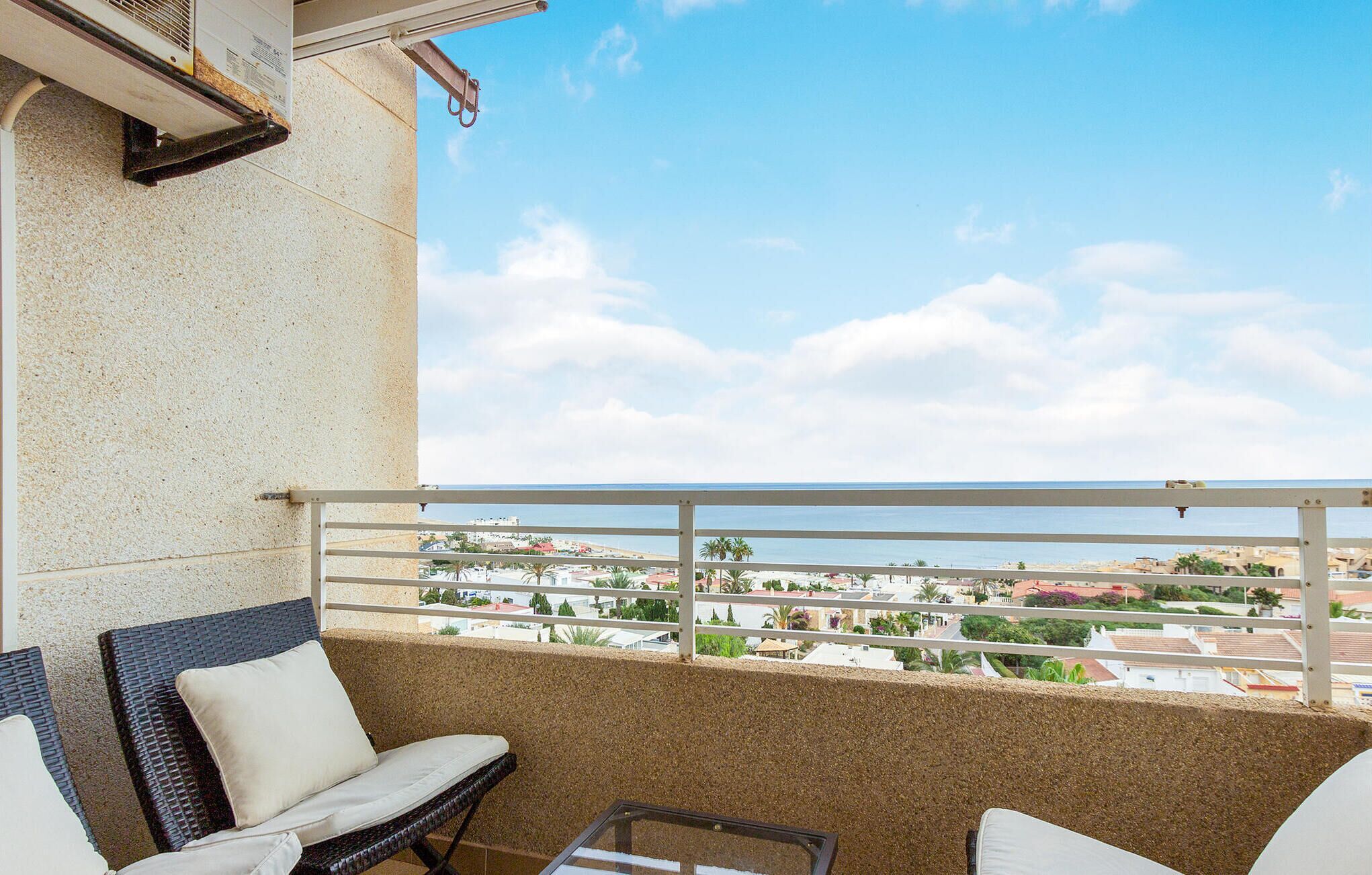 A balcony with wicker chairs and a glass door, overlooking a coastal town with buildings and a beach.