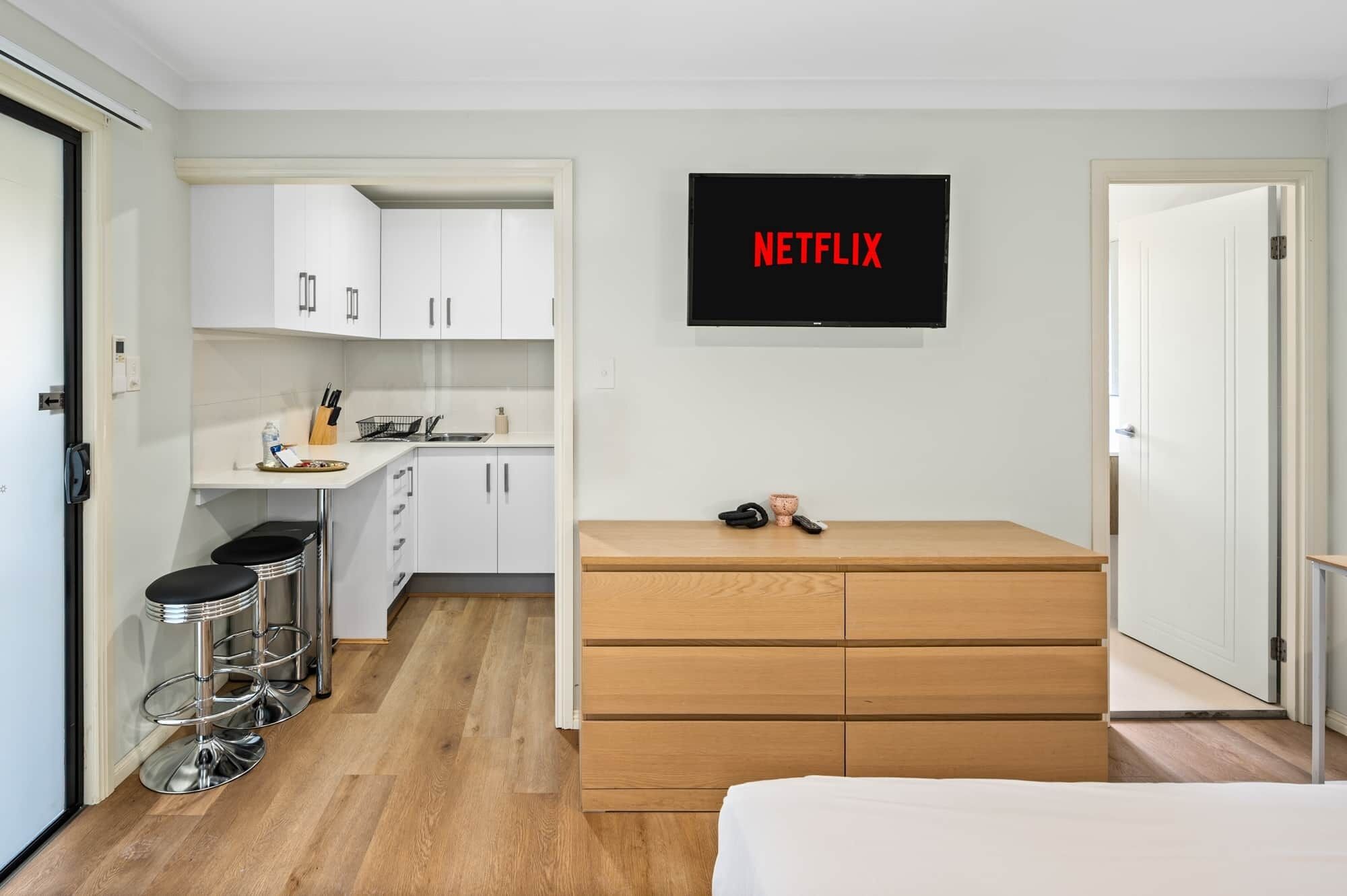 A compact room with a kitchenette, a bed, and a TV mounted on the wall displaying "NETFLIX".