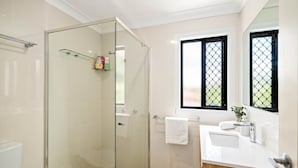 A modern bathroom with a glass-enclosed shower, a white vanity with a wooden cabinet, and a window with a black grid pattern. - Clean Sunnybank Studio | AC | Near Shops | 4144D0 (Sunnybank)