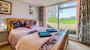 Un dormitorio con cama, una mesita de noche con lámpara, una ventana con cortinas y vistas a un campo verde.