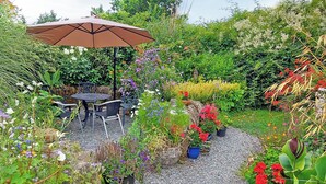 Un espacio al aire libre con mesa y sillas bajo un paraguas grande, rodeado de plantas y flores en macetas.