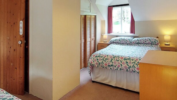 Un dormitorio con cama, puerta de madera, ventana con cortinas y una lámpara en una mesita de noche.