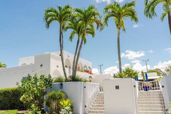 A white building with a staircase leading to a balcony, surrounded by palm trees and greenery.