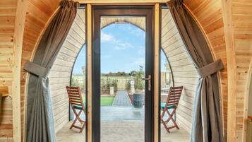 Une cabane en bois dotée d’une grande porte d’entrée en arc de cercle, de deux chaises en bois et offrant une vue sur un jardin.