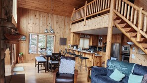 Interior d'una cabana de fusta amb menjador, cuina i saló que inclou un sofà i una escala.