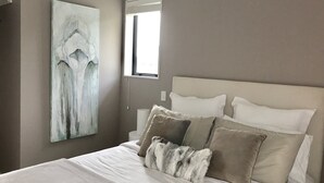 A bedroom with a large bed, a headboard, and a painting on the wall. - Villa Blanca in Palm Beach (Waiheke Island)
