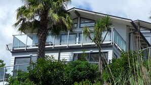 A modern multi-story building with large windows and balconies, surrounded by lush greenery. - Villa Blanca in Palm Beach (Waiheke Island)