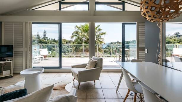 A spacious living area with a large window, a dining table, and a comfortable chair. - Villa Blanca in Palm Beach (Waiheke Island)