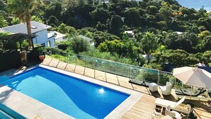 A swimming pool with a wooden deck, lounge chairs, and a glass railing overlooking a forested area. - Villa Blanca in Palm Beach (Waiheke Island)