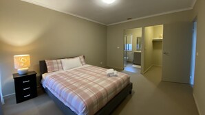 A bedroom with a bed, bedside table, and a lamp. There is a bathroom visible through an open door. - Whole House with 4 Queen Beds - Closer to Melbourne Airport - 8 Guests (Kurunjang)