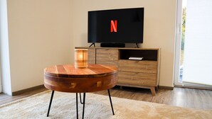 A modern living room with a wooden coffee table, a flat-screen TV displaying the Netflix logo, a wooden cabinet, and a large window.