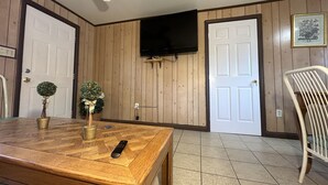 Una habitación con piso de madera, una mesa de comedor con un control remoto, dos plantas en macetas, un ventilador de techo y un televisor montado en la pared.