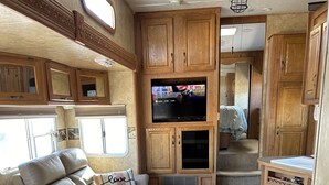 A well-lit RV interior with a sofa, a TV mounted on a wooden cabinet, and a kitchen area with a sink and cabinets.