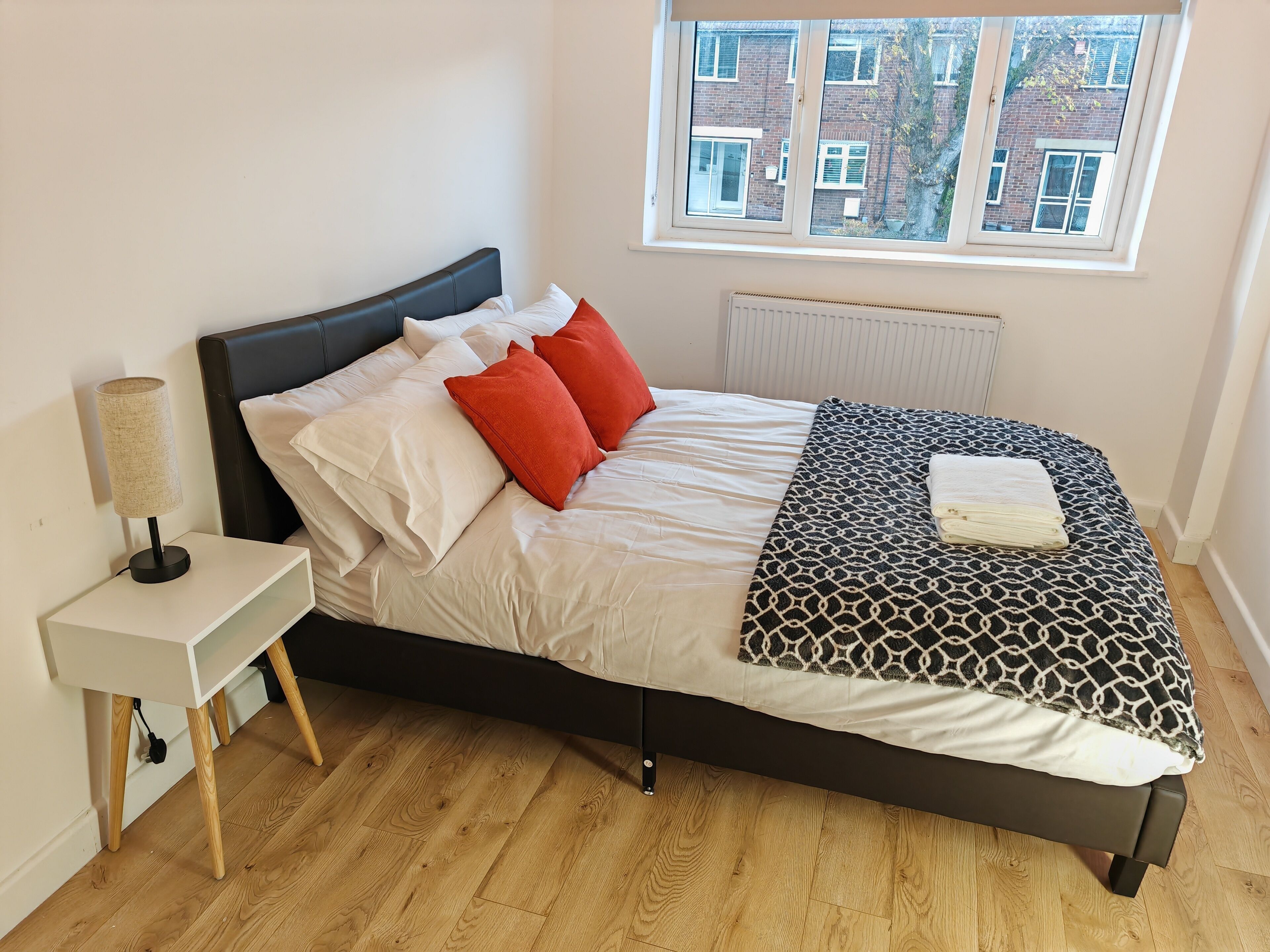A neatly made bed with white and red pillows, a patterned blanket, and a bedside lamp on a white nightstand.