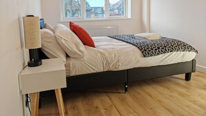 A bedroom with a bed, a nightstand, a lamp, a window with a view of houses, and a radiator. - Charming 5 Bedroom House in Bexley (Bexley)