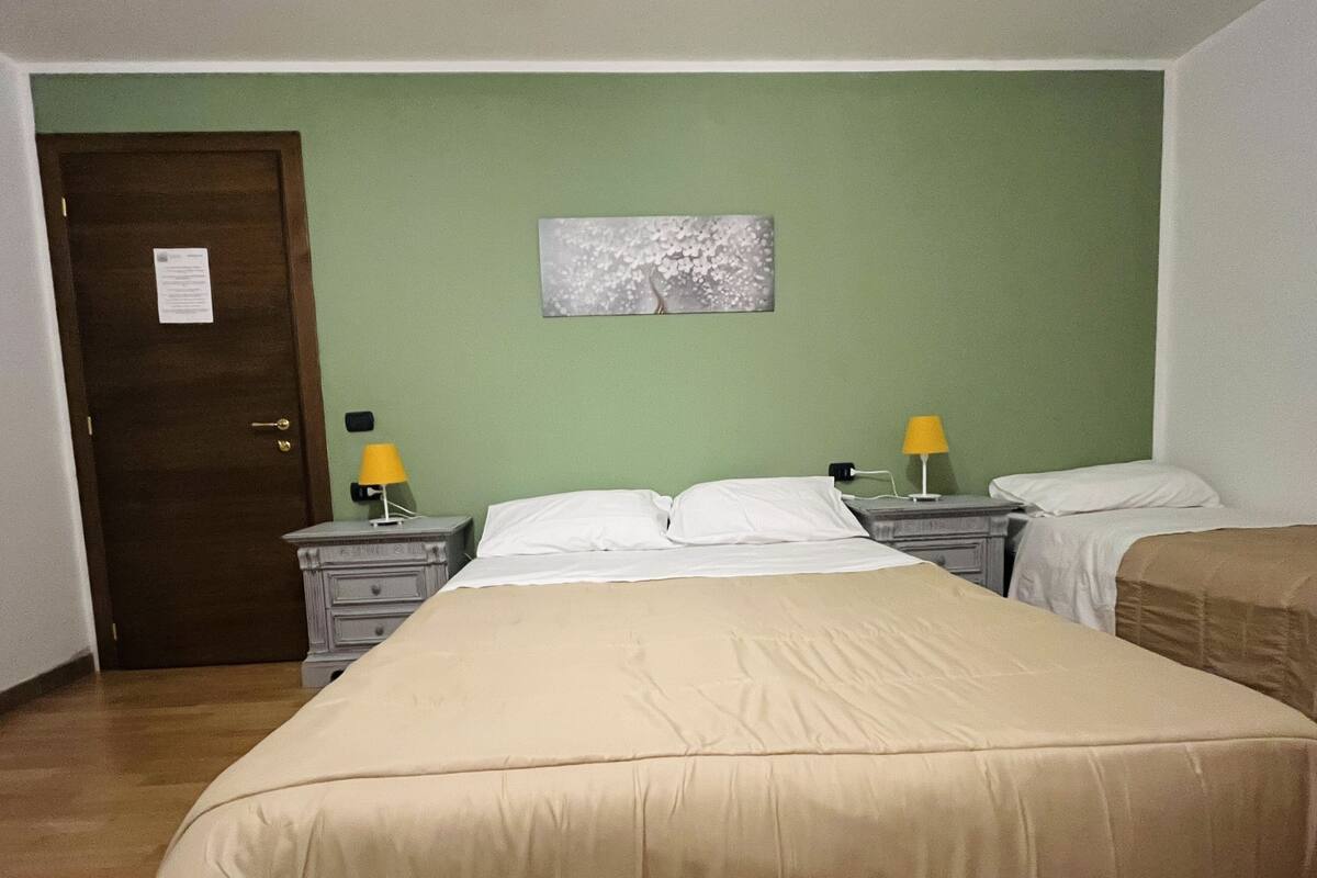 A hotel room with a bed, two bedside tables, a nightstand, a lamp, a door, and a wall-mounted picture.