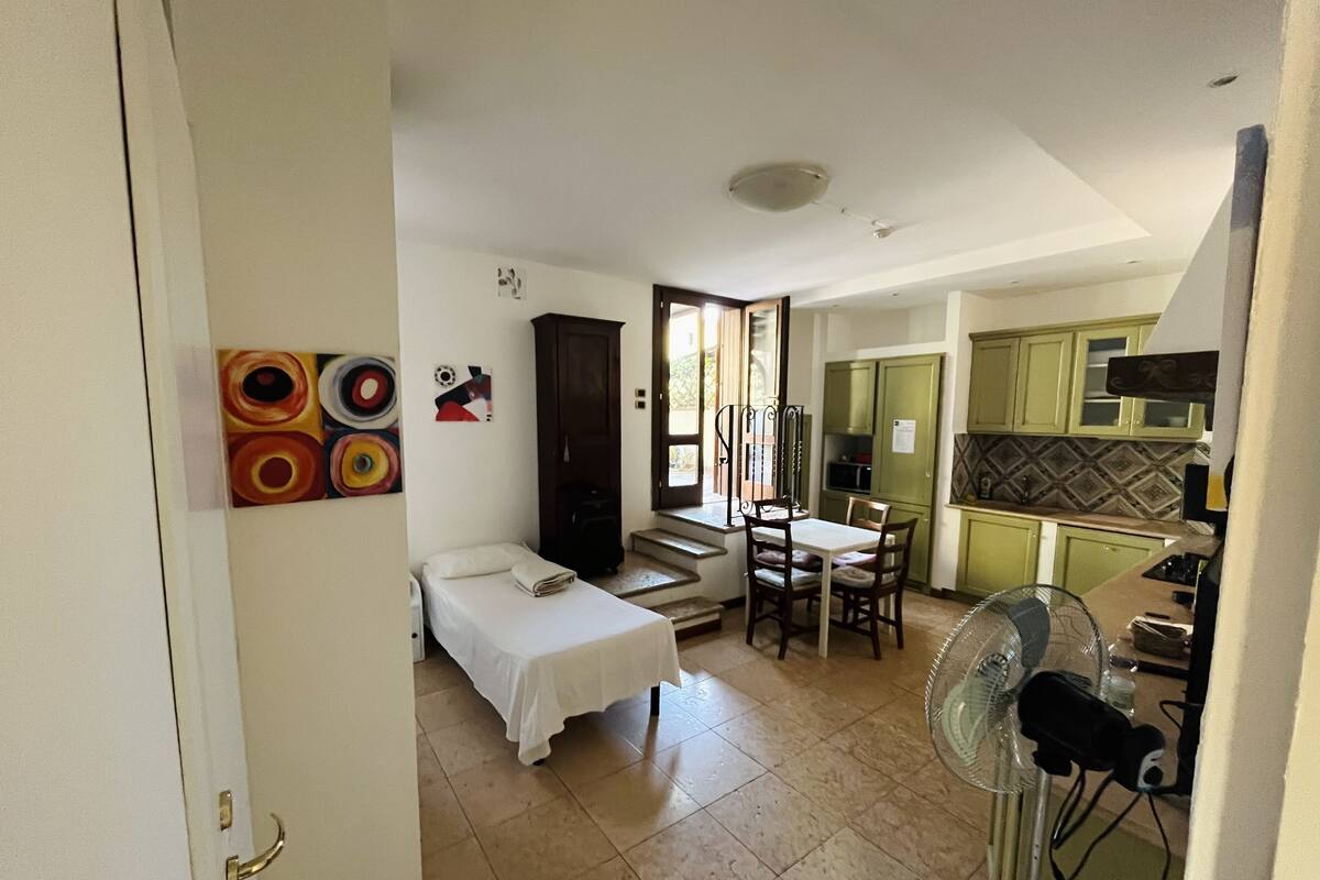A compact hotel room with a bed, a small table, and a fan. There is a kitchenette with green cabinets and a tiled backsplash.