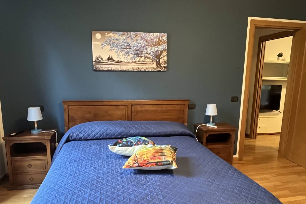 A bedroom with a large bed, two bedside tables, a nightstand, a wooden headboard, a wall-mounted painting, and a door leading to another room.