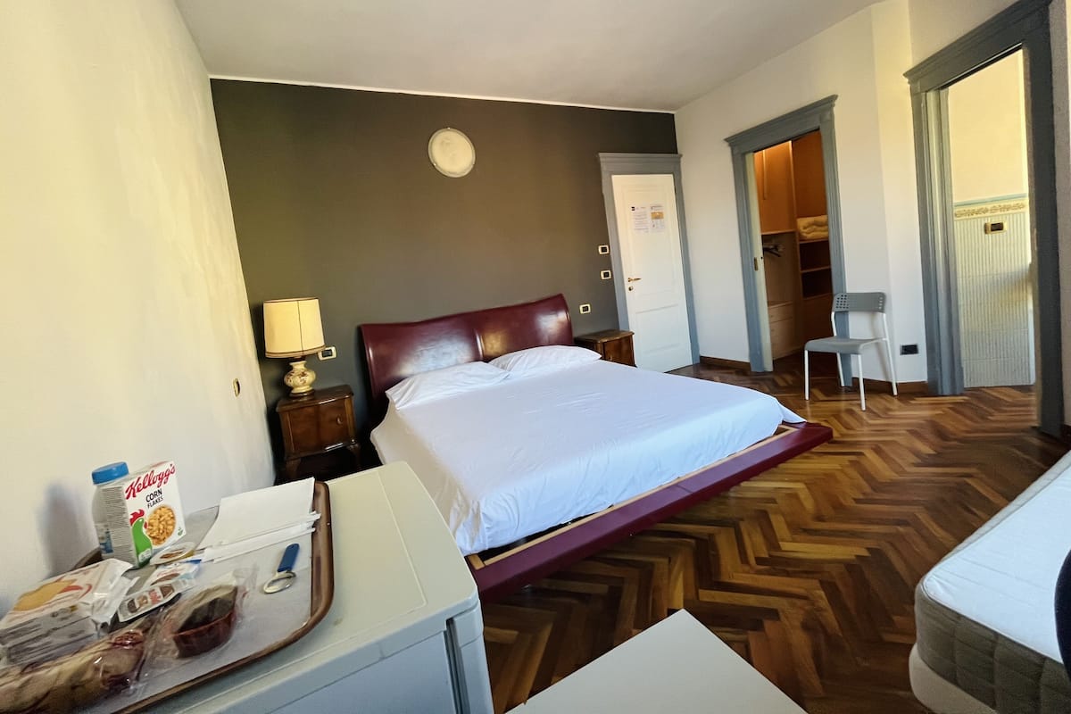 A hotel room with a bed, a desk with a lamp, a chair, and a kitchenette area.