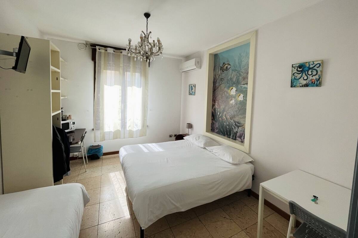 A hotel room with a bed, a desk, a chair, a chandelier, and artwork on the wall.