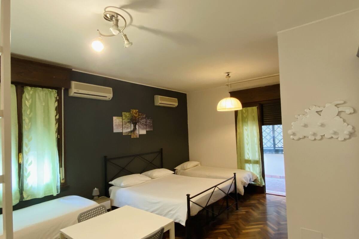 A hotel room with two beds, a table, and a chair. There are air conditioning units on the wall, a ceiling light, and a window with curtains.