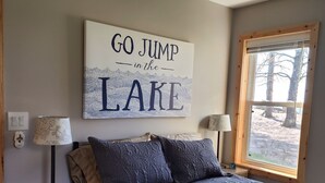 A bedroom with a bed, a nightstand, a lamp, a wall art piece, a window, and a ceiling fan. - Cozy cottage with private Lake Superior beach in Michigan’s Upper Peninsula  (Ontonagon)