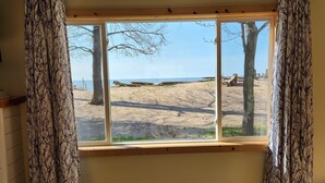 A room with a large window, wooden floor, and a view of a beach with trees and a clear sky. - Cozy cottage with private Lake Superior beach in Michigan’s Upper Peninsula  (Ontonagon)