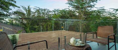 A balcony with wicker furniture, a tray with teapot and cups, and a view of trees and buildings.
