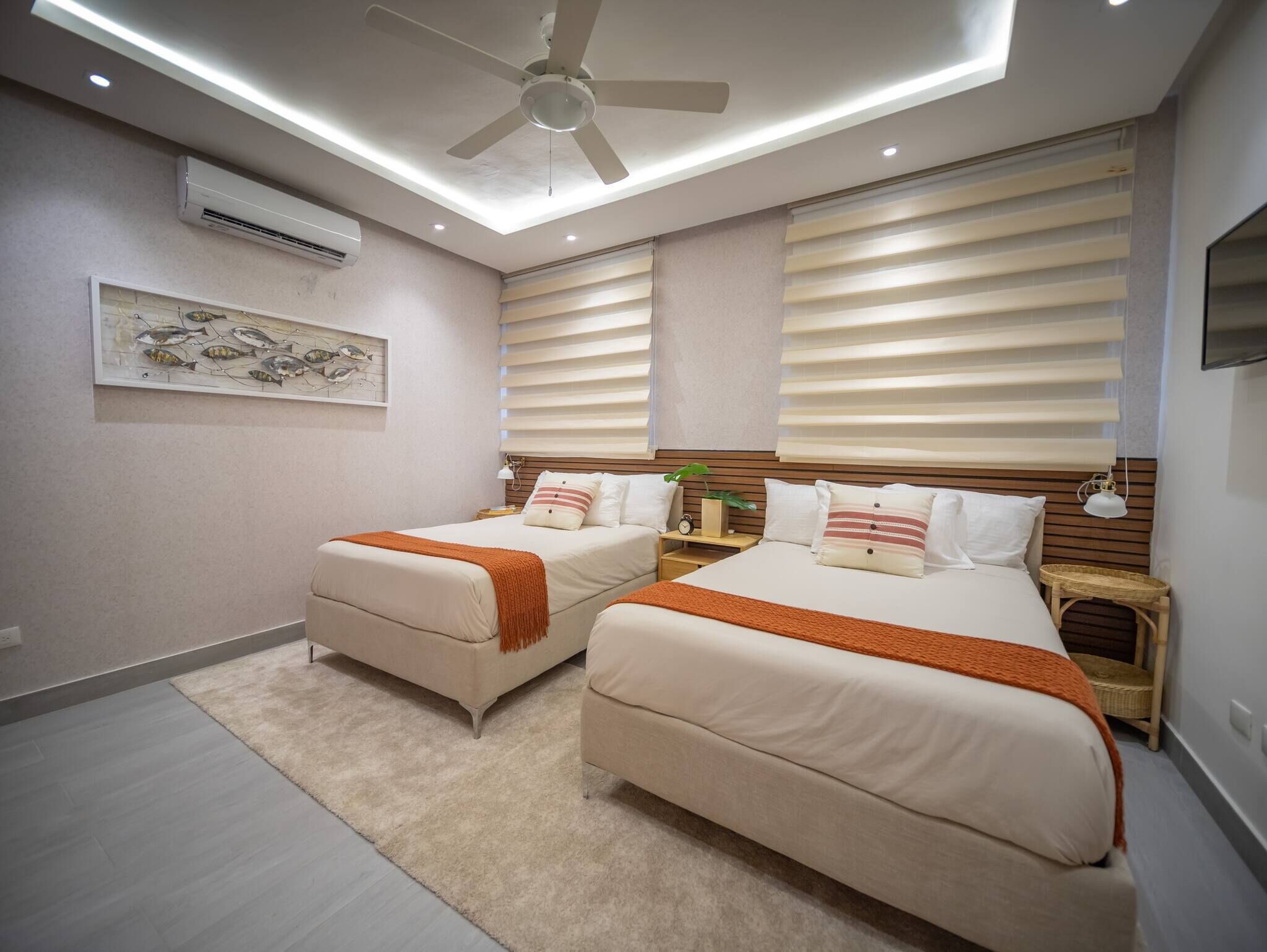 Un dormitorio con dos camas, ventilador de techo, iluminación empotrada, aire acondicionado instalado en la pared y una obra de arte con peces en un marco.