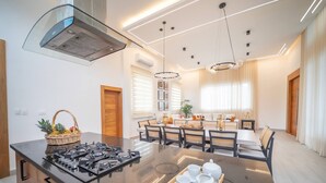 Una cocina moderna con una isla central, un comedor con lámpara de araña y un ventanal con persianas.