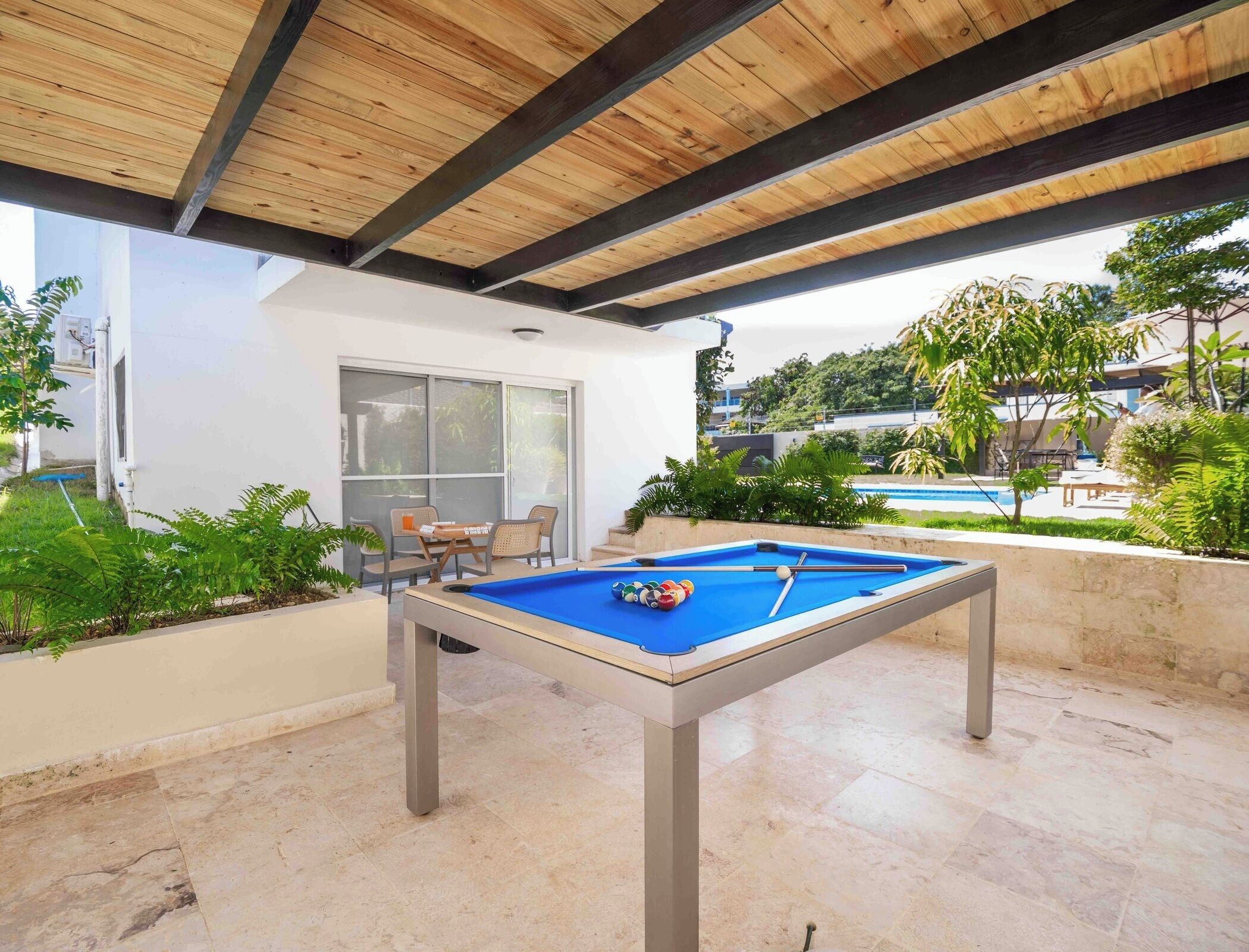 An outdoor pool table with a blue felt surface, surrounded by a patio area with a tiled floor.