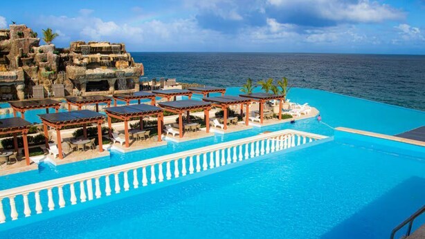 Una piscina de resort con vista despejada al océano, una estructura rocosa y varios espacios de estar cubiertos.