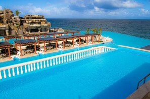 Ein Hotelpool mit freiem Blick auf den Ozean, einer Felsformation und mehreren überdachten Sitzbereichen.