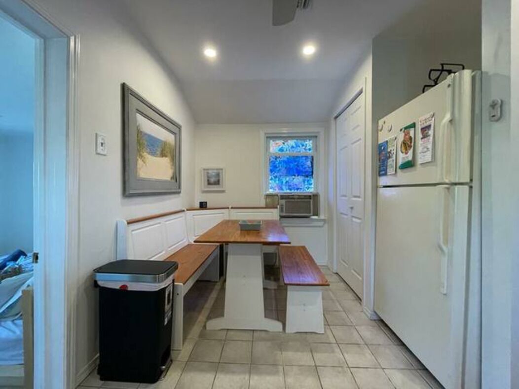 A narrow kitchen with a wooden table, white cabinets, a refrigerator, and a window with blue-tinted glass.