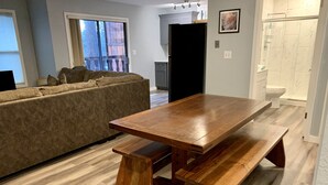 A modern living room with a wooden dining table and benches, a grey sofa, and a large window with a view of the outdoors. - Kennedy’s Camelot, a walkable family condo in beautiful Angel Fire! (Angel Fire)