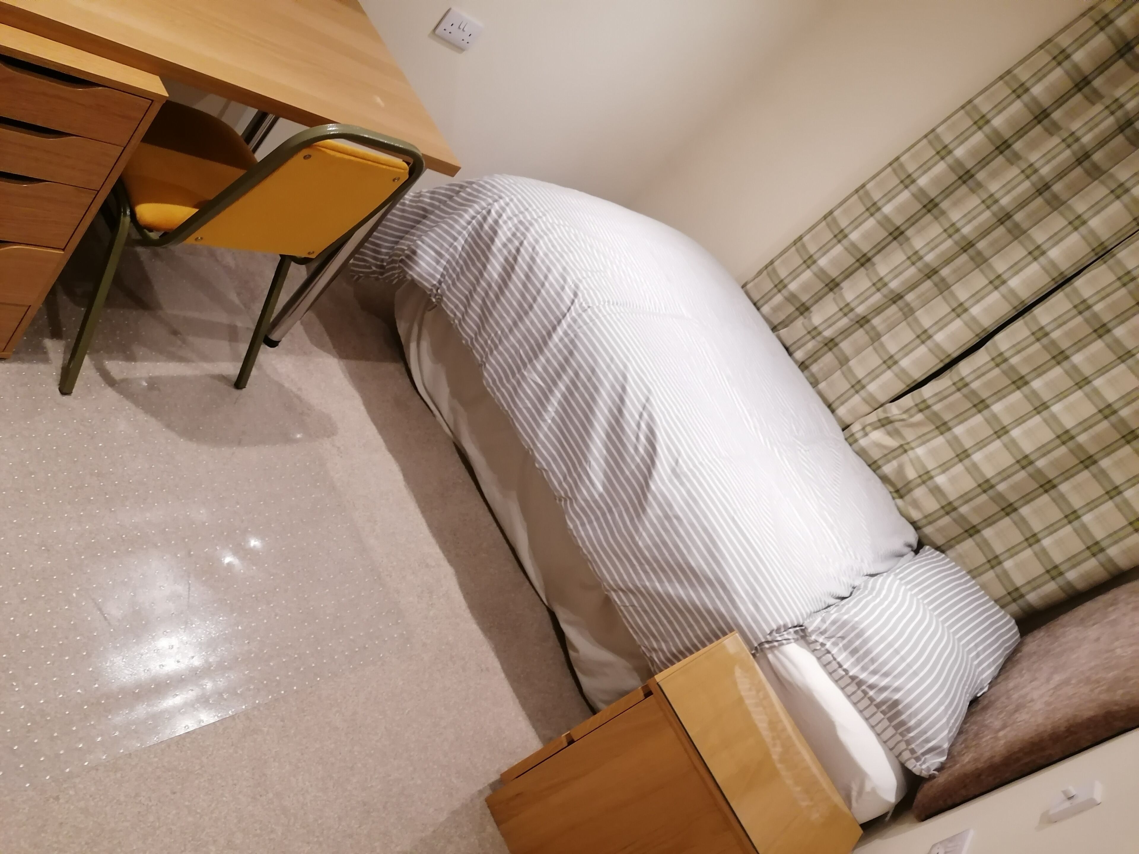 Et værelse med et træskrivebord, en stol, en seng med stribet sengetøj og en sofa med ternet mønster.