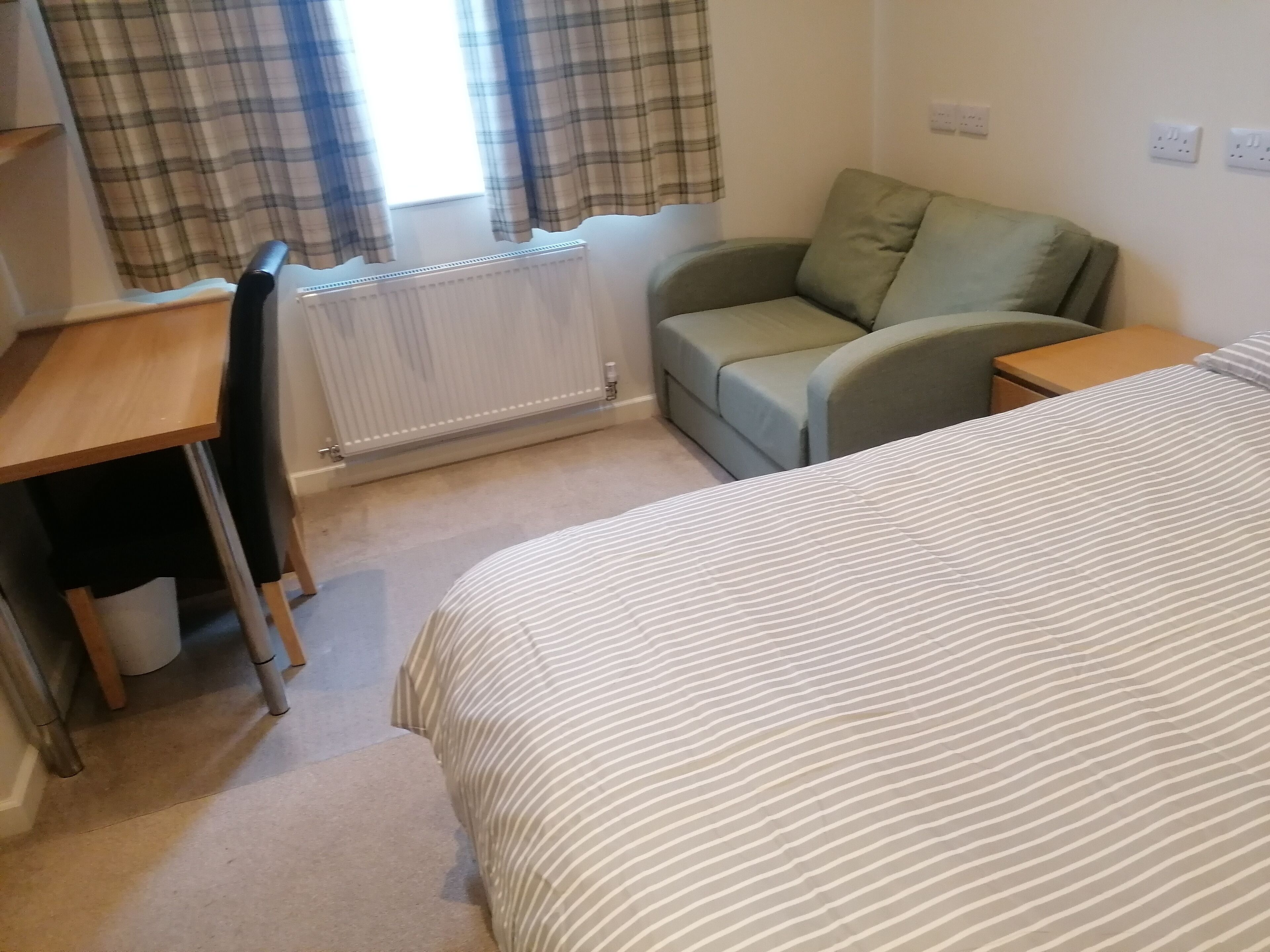 A bedroom with a bed, a desk, a chair, a sofa, and a radiator.