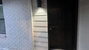Un mur extérieur en briques blanches, une porte noire et un luminaire vertical.