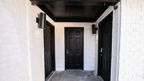 Un couloir étroit, avec une porte noire au fond, bordé de murs en briques blanches et surmonté d’un plafond noir.