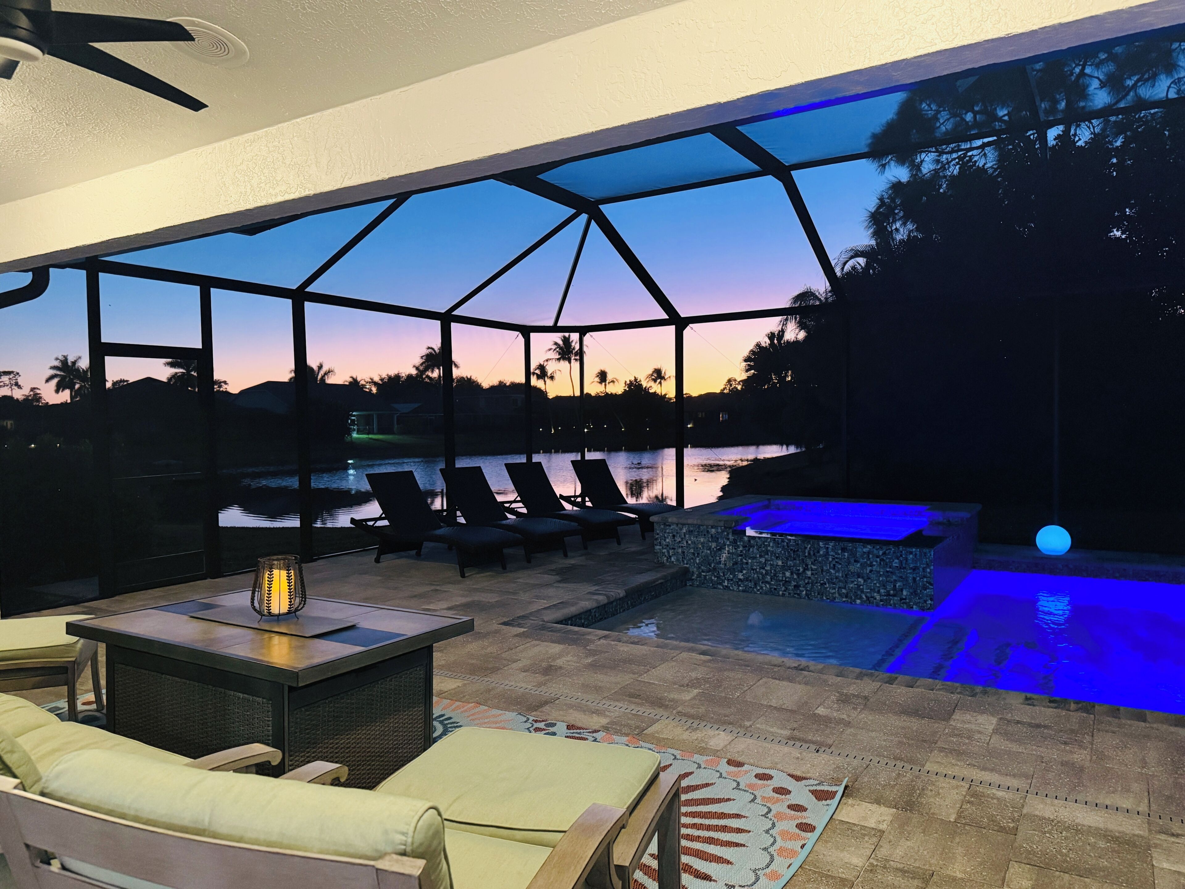 A covered outdoor seating area with lounge chairs and a table, overlooking a swimming pool and a body of water with a sunset view.