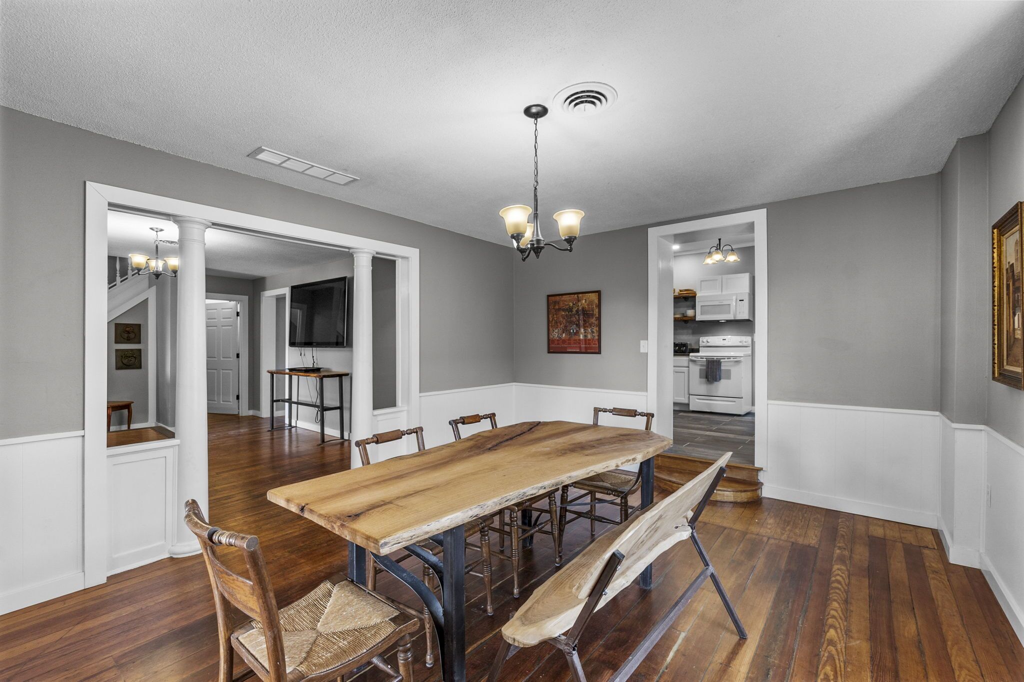 A dining room with a wooden table and chairs, a chandelier, and a view of the kitchen and living areas.