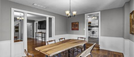 A dining room with a wooden table and chairs, a chandelier, and a view of the kitchen and living areas.