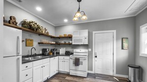 Una cocina moderna con armarios blancos, un fregadero de acero inoxidable, un microondas y una estufa. Se observa una repisa de madera con plantas y vajilla, así como un cubo de basura.