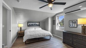 Un dormitorio con cama, mesita de noche, lámpara, cómoda, ventilador de techo y ventana con vistas.