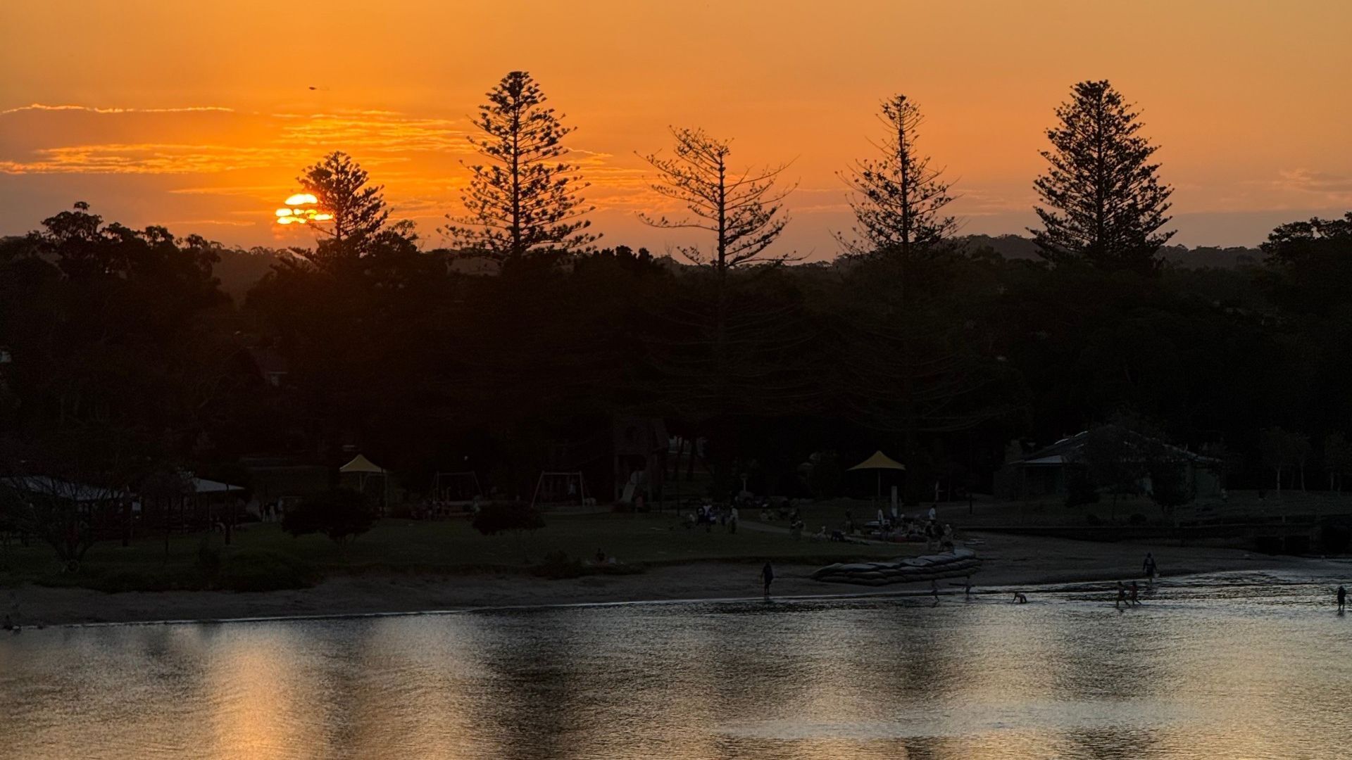 Un coucher de soleil sur une étendue d’eau, avec des arbres et quelques bâtiments en arrière-plan.