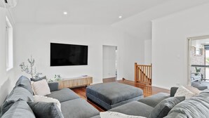 Ein modernes Wohnzimmer mit einem großen Flachbildfernseher, grauen Sofas, Holzböden und einer Treppe, die zu einem Außenbereich führt.