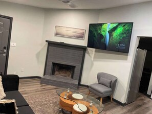 A living room with a fireplace, a flat-screen TV mounted above it, a glass coffee table, and a gray armchair.